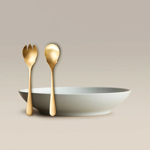 Two gold utensils a three-pronged fork and a spoon leaning against a white shallow bowl on a beige background