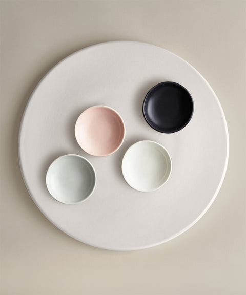 Five small bowls in pink, black, pale blue, white and light gray arranged on a round pale plate