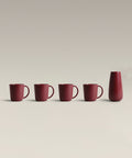 Row of four red mugs and a tall red bottle on a pale surface.