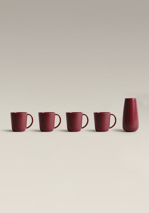 Row of four red mugs and a tall red bottle on a pale surface.